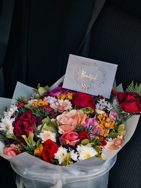 A vibrant bouquet of fresh flowers arranged in a round composition, placed in a clear wrapping with white tissue paper visible at the edges. The bouquet includes deep red roses, light pink lisianthus,