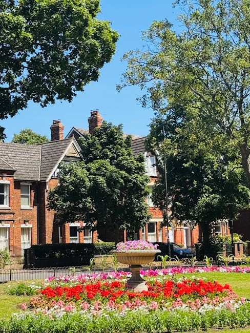 A clear view of a landscaped park with a central flowerbed filled with vibrant red, pink, and white blossoms, surrounded by well-maintained green grass and a decorative stone fountain on a raised plin