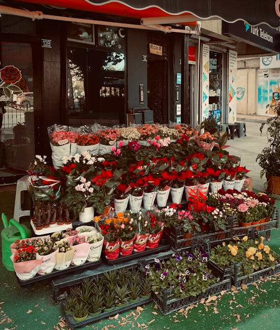 An outdoor display of fresh flowers in front of a shop on Station Road, featuring a variety of vibrant bouquets arranged on black plastic tables and plant racks. There are red and pink roses, white an